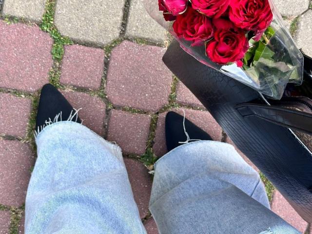 Barberry's legs in ripped gray jeans, black pointed shoes, standing on red brick pavement, holding a bouquet of red roses with green leaves.