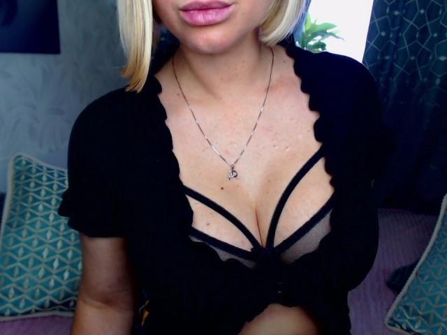 BelleLisaG with blonde hair, black ruffled top, and strappy black bra showing her tits, wearing a silver necklace with a heart pendant. Her lips are pink.