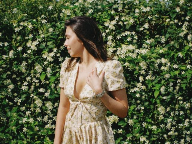 LounaLove has medium tits, wearing a floral dress with a deep V-neck, standing against a lush green bush with white flowers, looking to the side, hand near chest.