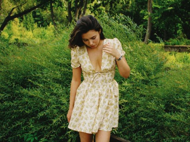 LounaLove with small tits, wearing a yellow floral dress with a deep V-neck, standing in green bushy foliage, looking down with wavy brown hair and a wristwatch.
