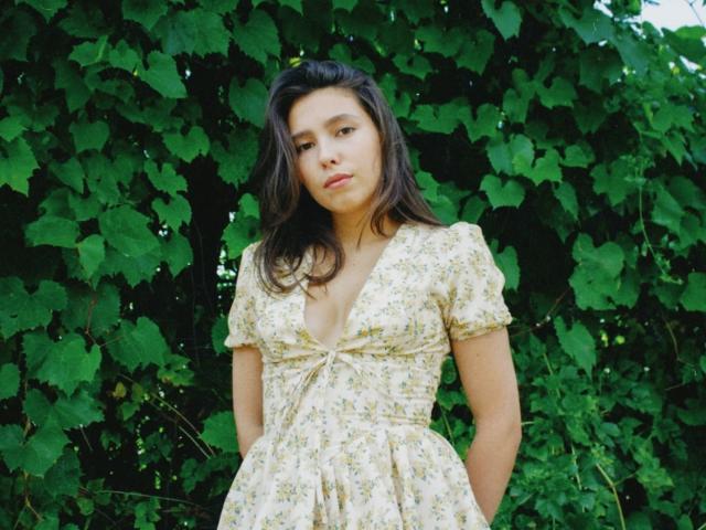 LounaLove with medium tits, wearing a floral dress, standing in front of green leafy vines, looking down with a neutral expression, brown wavy hair, fair skin.