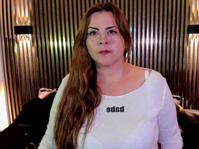AstridBayon ha lunghi capelli castani, una camicia bianca e una collana, ed è in piedi con un'espressione neutra. Ha un seno grande e morbido e un viso carino, leggermente paffuto..