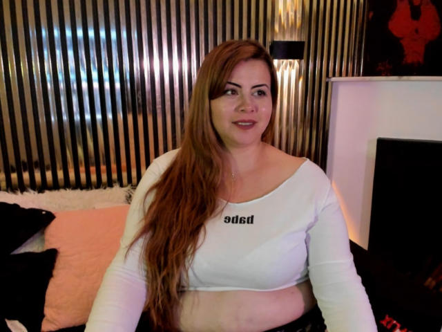 Astrid Bayon, com cabelos vermelhos longos, blusa branca de manga comprida, mostrando seus seios, sentada, sorrindo, pele clara, em um quarto com parede listrada em preto e dourado..