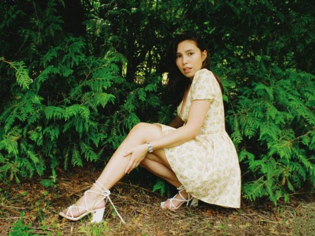 LounaLove crouching, wearing a floral yellow dress, white strappy heels, bracelet, and looking at the camera with a slight smile. She's surrounded by green foliage.