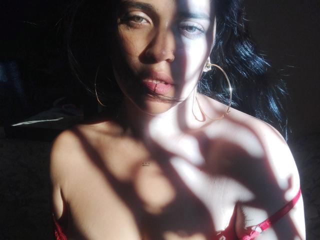 AgathaK with dark hair, large hoop earrings, and red bra straps, sunlight and shadow on her face and tits, slightly parted lips, soft light on her brown skin.