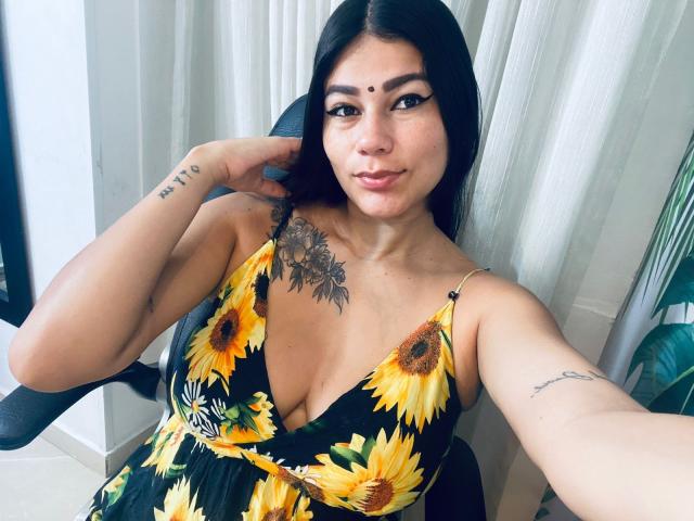 KeraniMarala met een jurk met zonnebloemen, middelgrote borsten, tatoeages op de borst en arm, donker haar, bruine huid, zittend in een stoel, kijkend naar de camera, een kleine zwarte stip op het voorhoofd..