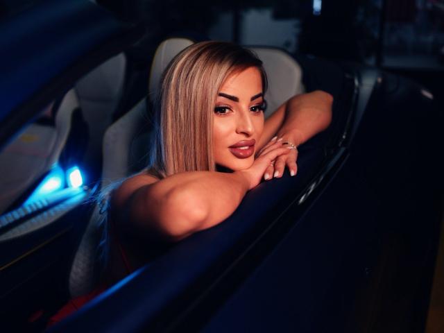 MissEmaDivine with blonde hair, pouty lips, and dark eyes leans on a car's blue interior, her arms resting on the top, looking seductive in low light.