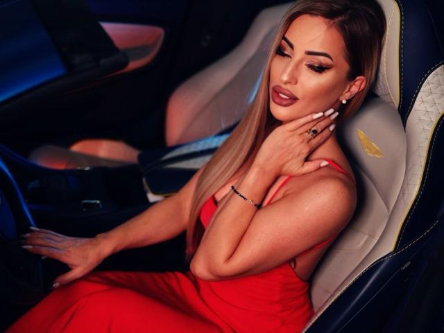 MissEmaDivine with long blonde hair, wearing a red dress, sitting in a car, hand on neck, looking down, white nails, ring, bracelet, and a sexy, confident expression.
