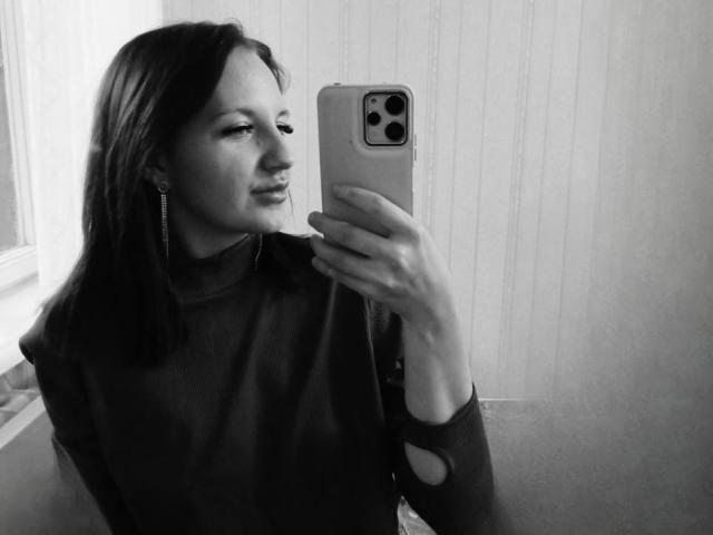 RosaNelsson with shoulder-length dark hair, wearing a dark sweater, taking a selfie with an iPhone, showing off her small ass in a black-and-white photo.