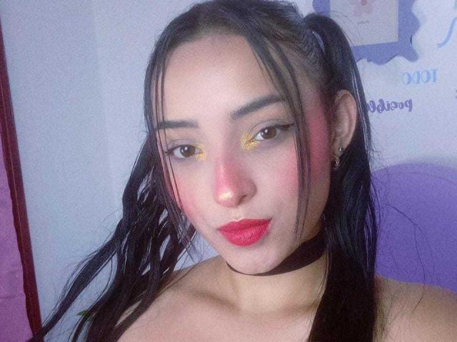 MaraLewis has long black pigtails, yellow eyeshadow, red lipstick, and a black choker. Her brown eyes and light skin are visible. She's looking at the camera with a slight smile.