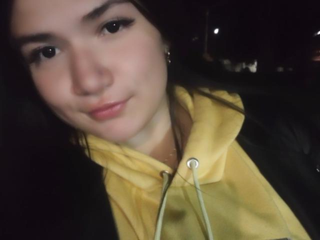 AmyRousse, avec une peau claire, des sourcils noirs et des lèvres roses, portant un sweat à capuche jaune avec une veste noire, des cheveux foncés, un sourire discret et de petites boucles d'oreilles, dans un environnement peu éclairé..
