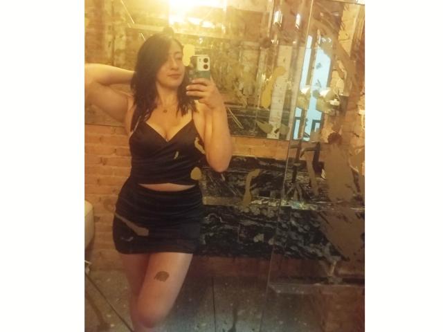 ZaraBeicker with wavy dark hair, black strapless top, and black skirt, showing her tits and ass in a mirror selfie, standing in a dimly lit, rustic room.