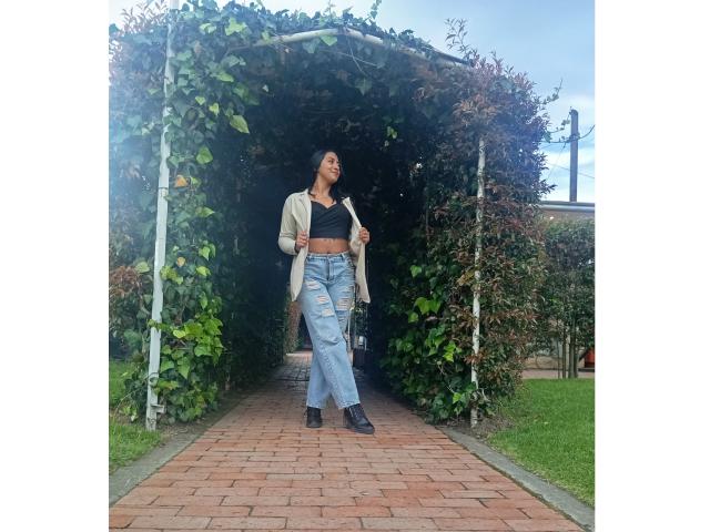 ZaraBeicker with a slim build, dark skin, and medium tits, wearing a black crop top, light jacket, and ripped jeans, standing under a green leafy archway, looking up.