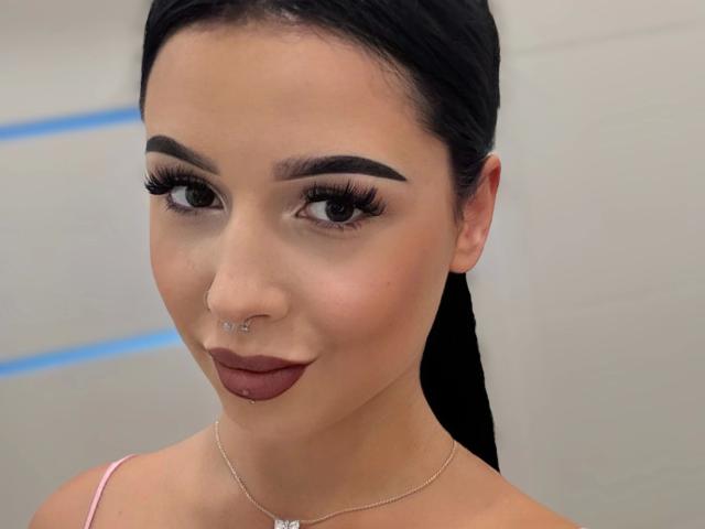 TaliaClassy-hot has dark hair, thick eyebrows, long lashes, a nose ring, and brown lipstick. She's wearing a pink strap and a silver necklace. Her ass is not visible.