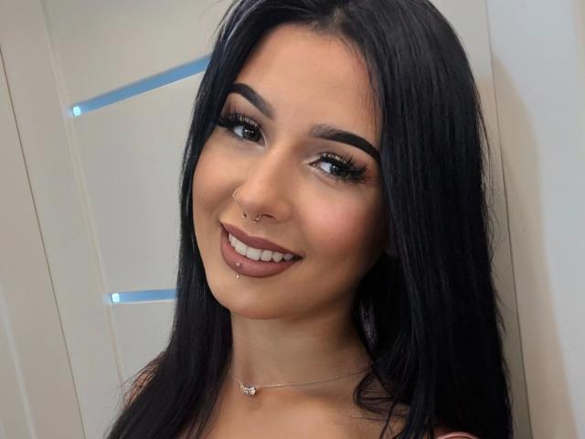 TaliaClassy-hot has long black hair, brown eyes, thick eyebrows, a nose piercing, a lip piercing, and is smiling with her tits slightly visible, wearing a silver necklace.