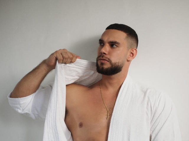 ChrisBlake with a short beard, white towel open to reveal a shirtless chest, gold cross necklace, looking to the side, holding towel with right hand.
