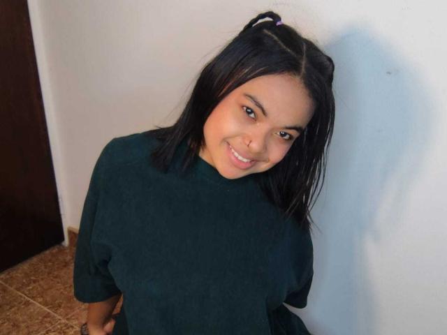 SoleneFontaine with black hair, green t-shirt, smiling, nose ring, and ponytail, leaning against a white wall, brown tiled floor, and dark door in the background.