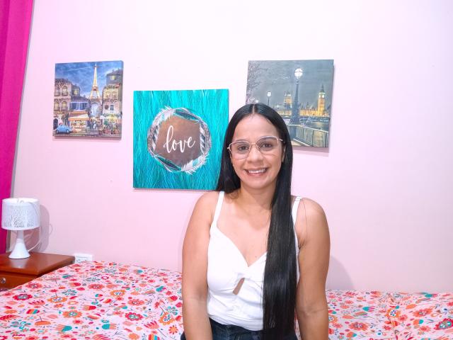 ThreeSensations has long black hair, glasses, and a white tank top showing her small tits, sitting on a bed with a floral pattern, smiling. Pink wall and paintings.