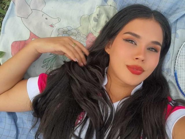 CarolineWill has long black hair, red lipstick, and is wearing a sports shirt with red and white sleeves, lying on a bed with a floral pillow, touching her hair.