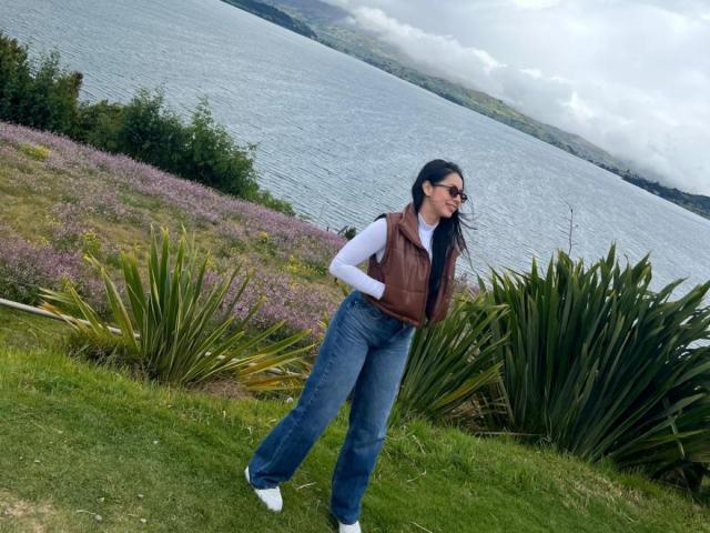 IsabellaJames with long black hair, wearing sunglasses, brown vest, white shirt, blue jeans, and white shoes, standing on grassy hill with purple flowers and green plants.