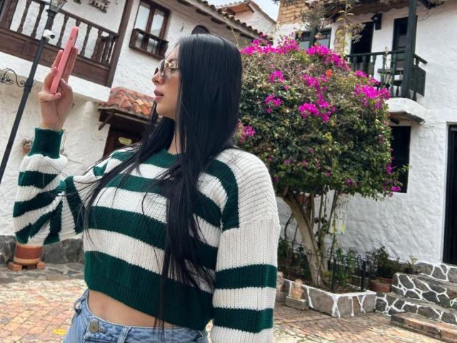 IsabellaJames with long black hair, wearing green and white striped sweater, holding pink phone, showing her tits, standing in front of white house with purple flowers, wearing glasses.