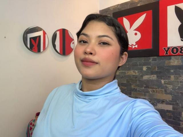 ShailaYojana with short black hair, light skin, and pink lips, wearing a light blue turtleneck, smiling slightly, standing in front of red and black wall decor.