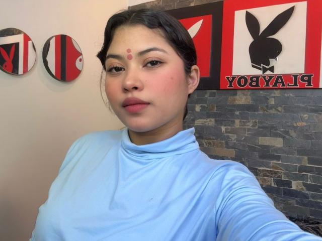 ShailaYojana with medium tits, wearing a light blue turtleneck, has a red dot on her forehead, black hair, and a neutral expression, in a room with Playboy decor.