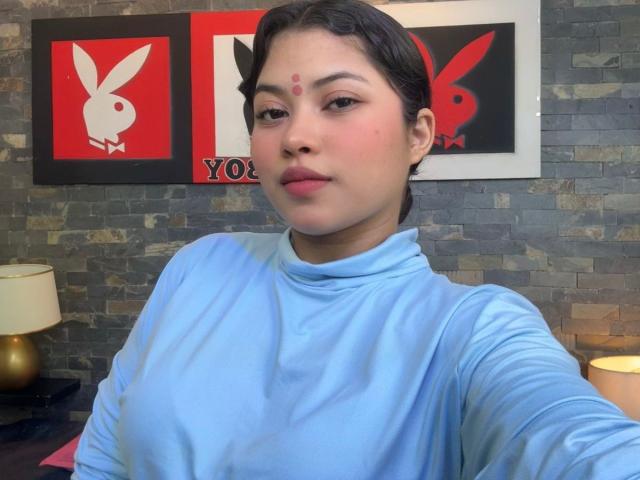 ShailaYojana, Asian woman with medium skin, dark hair in a bun, wearing a blue turtleneck, red dot on forehead, looking at camera, slightly smiling, with small, cute tits.