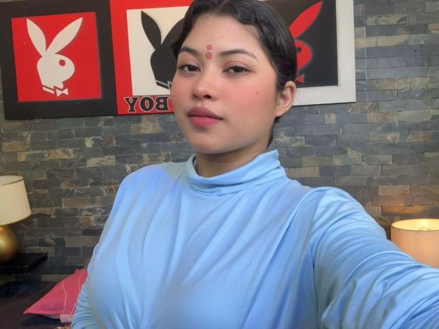 ShailaYojana with short black hair, light brown skin, red dot on forehead, wearing a blue turtleneck, small tits, in a room with stone wall and Playboy posters.