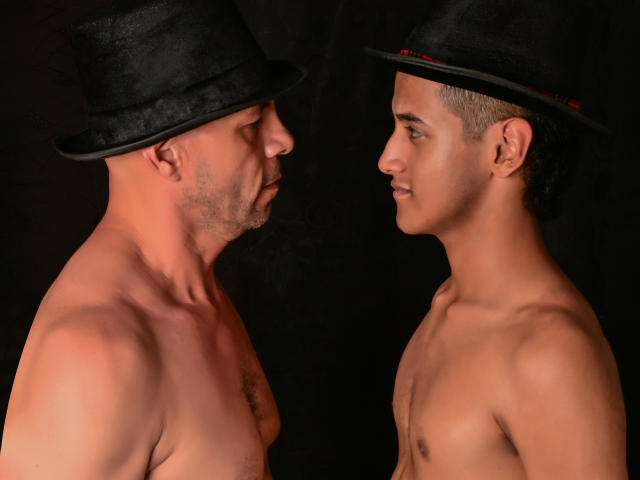 Two shirtless GroupFantasies with black hats, facing each other, showing off toned torsos and serious expressions against a dark background. One has a bald head, the other has short hair.