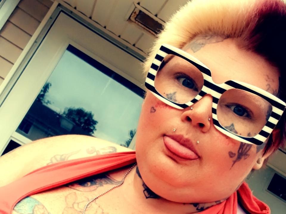 PandaPie with striped glasses, tongue out, has tattoos, piercings, short blonde hair with red tips, and a red top, showing off her tits and a choker.