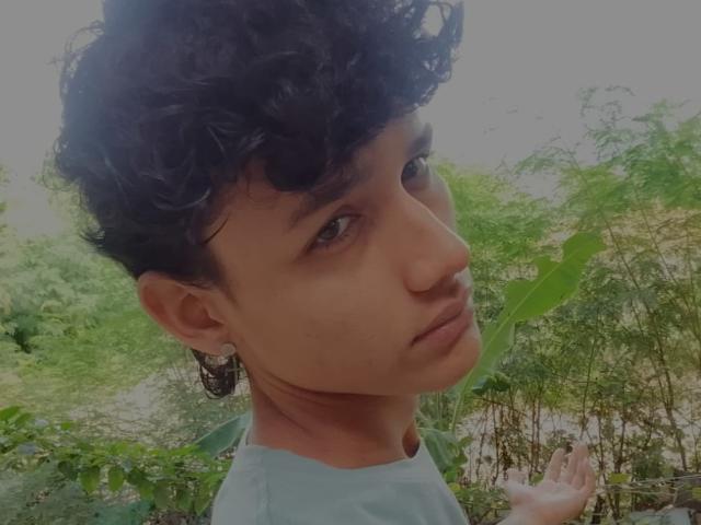 Curly-haired Alenders with brown skin and dark eyes, wearing a green shirt, looking over shoulder with a serious expression, has small butterfly earring on left side.