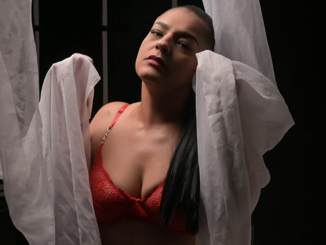 GroupFantasy with dark hair, red lace bra, and white semi-transparent cloth, looking up, showing her tits, in a dimly lit, dark background.