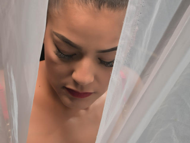 GroupFantasy with fair skin, dark eyebrows, red lipstick, and long eyelashes, looking down, partially hidden by white sheer curtains, her bare shoulders visible.