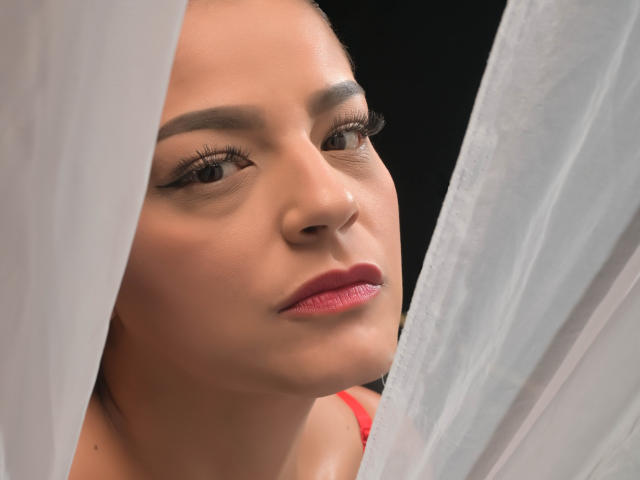 GroupFantasy's face, showing pink lipstick, subtle makeup, and eyeliner, peeking through white curtains, with soft light on her tan skin, brown eyes, and relaxed expression.