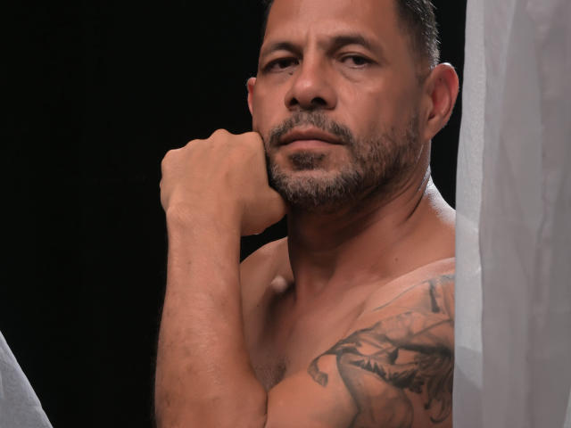 Shirtless GroupFantasy with tattoos, dark hair, and beard, resting chin on hand, looking serious, white curtain on right, black background.