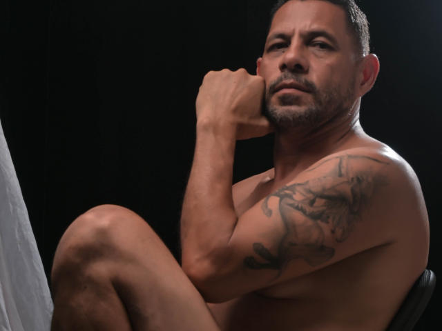 Shirtless GroupFantasy with a tattoo on his right arm, short dark hair, and a beard, sitting with his knee up and fist resting on his chin.