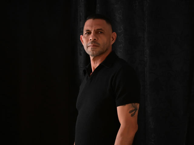 GroupFantasy is a muscular dude with short black hair, a black polo, and a tattoo on his right arm, standing against a dark curtain. He's looking serious.
