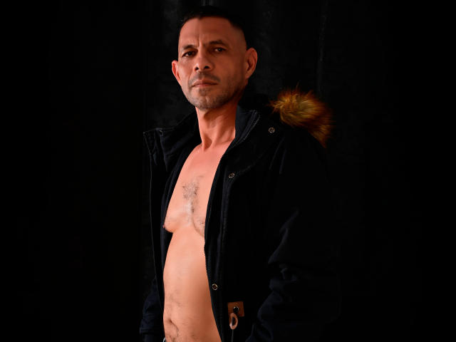 GroupFantasy with a muscular, hairy chest, wearing an open black jacket with a fur collar, looking serious, against a dark background. Short-cropped hair, slightly graying, and stubble.