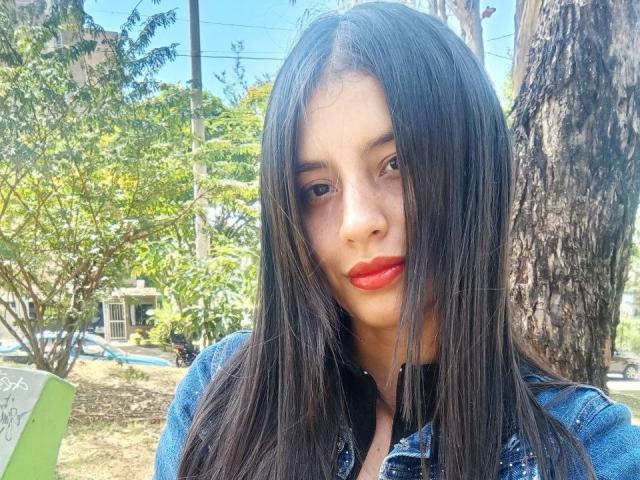 MadissonFontaine has long black hair, red lipstick, and a denim jacket. She's standing outside with a tree behind her, looking at the camera with a serious expression.