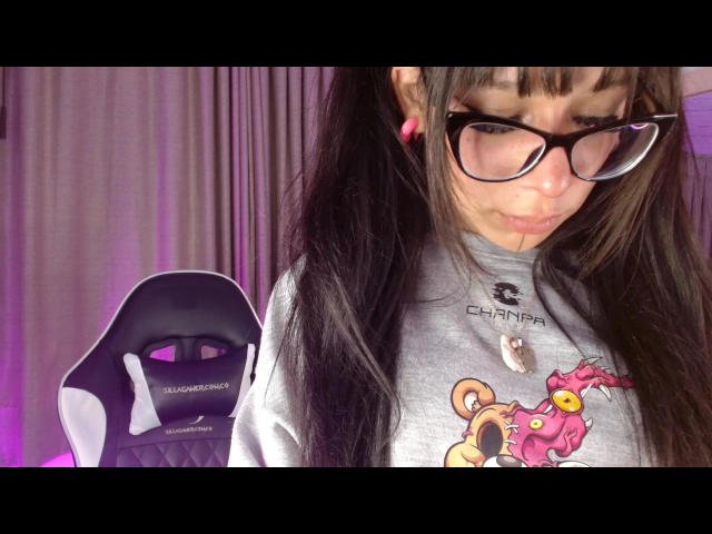 JenniPurple with long black hair, glasses, pink earrings, gray shirt, and cute cartoon print, sitting on a black and white gaming chair, looking down. Boobs are not visible.