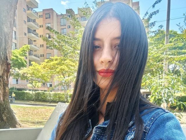 MadissonFontaine with long black hair, red lipstick, and blue denim jacket, looking down with a slight pout, sunny day, green trees, and apartment building in the background.