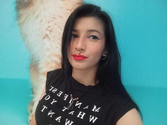 AlisKooper with black hair, red lipstick, nose ring, and black sleeveless top, standing next to a fluffy white and brown animal, against a blue background.