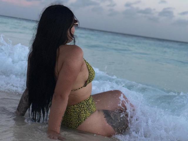 MalakCote, con cabello largo y negro, vestida con un bikini amarillo de estampado de leopardo, está sentada en aguas poco profundas del océano, de espaldas a la cámara, con olas alrededor de los muslos, un tatuaje en el muslo derecho, y un cielo crepuscular..