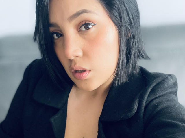 SamantaMuller has short black hair, brown eyes with winged eyeliner, and a pouty expression. She's wearing a black coat, looking directly at the camera with a slight blush.