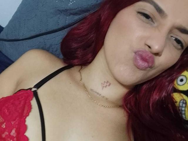 MiaHotGirll69 with red hair, pouty pink lips, black and red lace bra, gold necklace, tattoo below chin, lying down, looking at camera, slightly winking, cute.