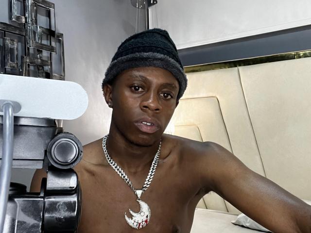 OriionBlack is shirtless, wearing a black beanie and a silver chain with a red diamond-studded pendant, looking serious with a camera in the foreground.