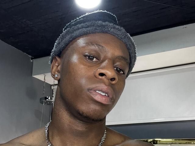 OriionBlack has a dark brown skin tone, short black hair in a fuzzy cap, and is wearing a chain necklace with small earrings. His face is slightly tilted upward.