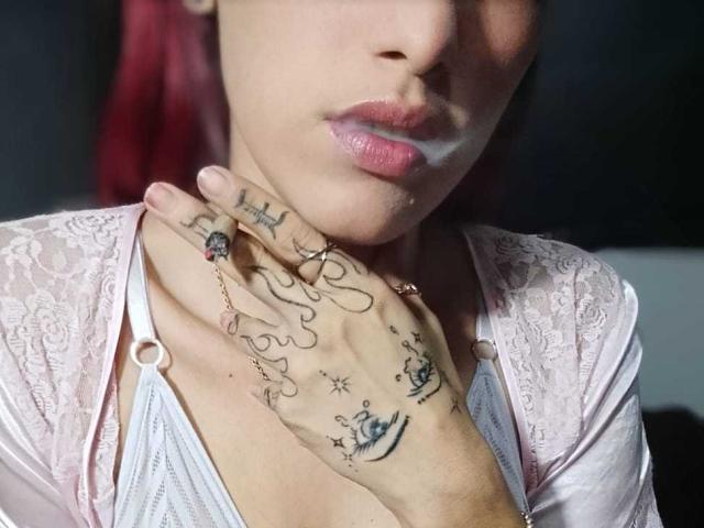 MiaHotGirll69's hand with tattoos on her fingers, pink lipstick, and red hair, wearing a lace pink top, touching her neck. Boobs visible in the white top.