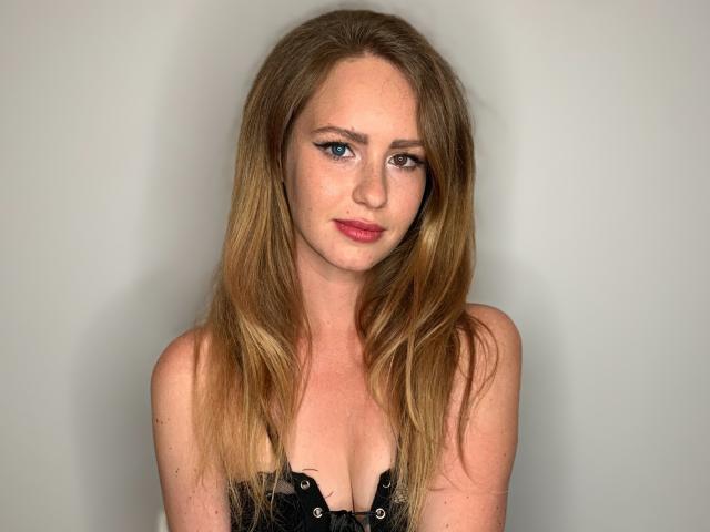LyraFantasy has blonde, wavy hair, blue eyes, and pink lipstick. She's wearing a black, strapless top with visible cleavage. Her expression is neutral, looking slightly to the right.